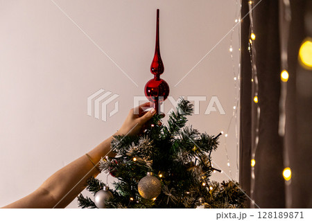 placing a shiny red christmas tree topper on a decorated artificial tree with warm string lights and festive ornaments in a cozy interior placing a shiny red christmas tree topper on a decorated artificial tree with warm string lights and festive ornaments in a cozy interior 128189871
