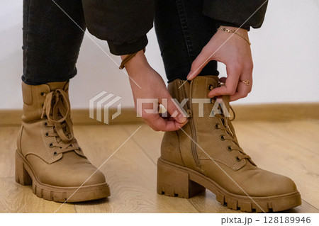 adjusting zipper on stylish brown leather boots with chunky heels and laces, close-up of hands and legs indoors on wooden floor 128189946