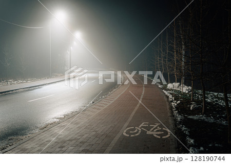 Empty road and bike path in a foggy nighttime setting, illuminated by streetlights. 128190744