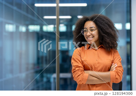 Young adult African American woman stands confidently with crossed arms in a modern office setting, smiling and looking away 128194300