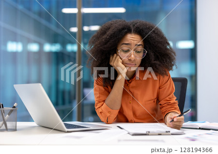 A woman with glasses sits at her desk, looking concerned while reviewing documents and a laptop. Her expression suggests she is stressed. A woman with glasses sits at her desk, looking concerned while reviewing documents and a laptop. Her expression suggests she is stressed. 128194366