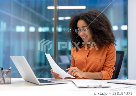 Successful businesswoman at workplace inside office working with documents, reports and contracts. Woman smiling happily, using laptop at work. 128194372