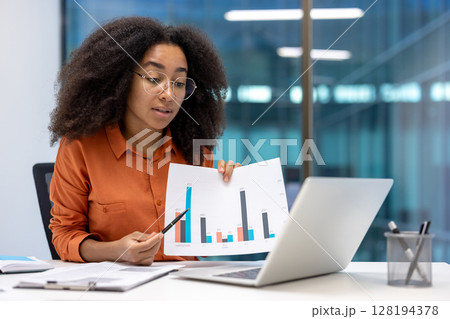 A focused woman presents a data chart to an audience via laptop during a video conference in a modern office setting. She holds a pen for emphasis. A focused woman presents a data chart to an audience via laptop during a video conference in a modern office setting. She holds a pen for emphasis. 128194378