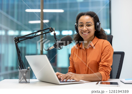 An African American woman smiles while working on a podcast with a microphone and laptop at a modern office. An African American woman smiles while working on a podcast with a microphone and laptop at a modern office. 128194389