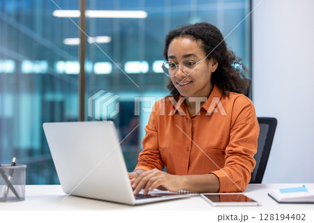 A young businesswoman works on her laptop in a modern office, smiling and focused. The scene shows a professional and productive environment. A young businesswoman works on her laptop in a modern office, smiling and focused. The scene shows a professional and productive environment. 128194402