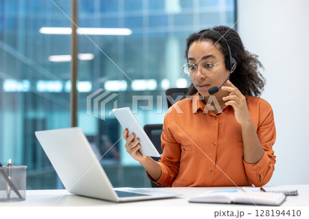 A woman with a headset and tablet works in a modern office, laptop visible nearby, engaging in customer service. 128194410