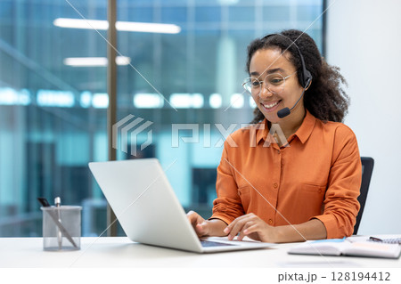 Smiling customer service representative wearing a headset and glasses working on a laptop in a modern office setting. Focus on communication and support. Smiling customer service representative wearing a headset and glasses working on a laptop in a modern office setting. Focus on communication and support. 128194412
