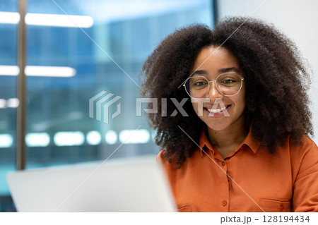 A beautiful young woman with curly hair and glasses smiles at the camera while working on a laptop in an office setting. 128194434