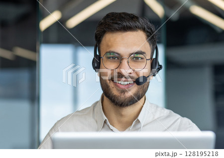 A smiling customer service representative wearing a headset, providing support. He is looking at the camera with a friendly expression in an office setting. A smiling customer service representative wearing a headset, providing support. He is looking at the camera with a friendly expression in an office setting. 128195218