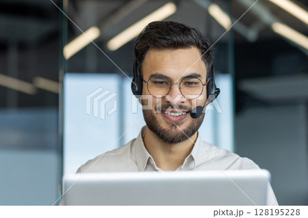 Man with headset close-up smiling, talking and consulting clients remotely. Support service worker at workplace inside office. Consultant at work. Man with headset close-up smiling, talking and consulting clients remotely. Support service worker at workplace inside office. Consultant at work. 128195228