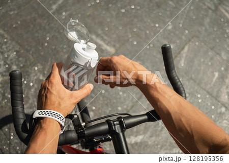 Close up of hands of cyclist holding water bottle, standing with his bike in park on sunny day. Man takes a break from his bike ride. Close up. 128195356
