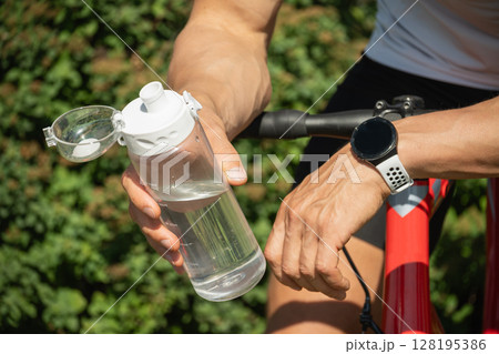 Close up of hands of cyclist holding water bottle, standing with his bike in park on sunny day in forest. Close up. 128195386