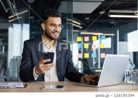 A smiling businessman in a suit is using a phone and laptop at his desk, possibly checking emails or communicating. 128195394