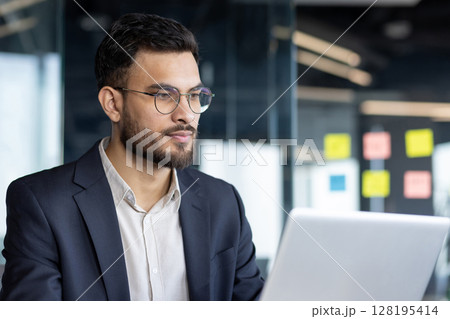 Close-up photo of serious confident businessman at work, thinking man reading electronic report. Businessman at workplace inside office working with computer. Close-up photo of serious confident businessman at work, thinking man reading electronic report. Businessman at workplace inside office working with computer. 128195414