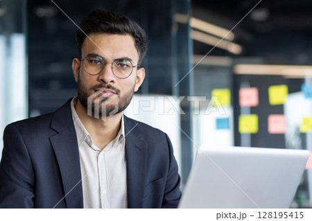 An entrepreneur is working on his laptop in a modern office. He has a determined look and wears a suit jacket over a dress shirt 128195415