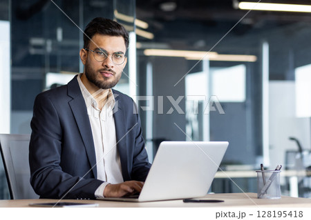 A confident businessman in a navy suit and glasses, working on a laptop in a modern office setting. 128195418