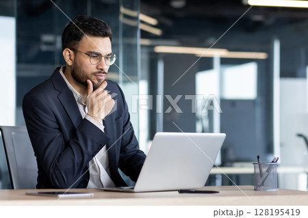 Businessman in dark suit analyzing work on laptop in office. He appears thoughtful and focused, embodying professionalism. Scene reflects modern corporate environment and digital productivity. 128195419