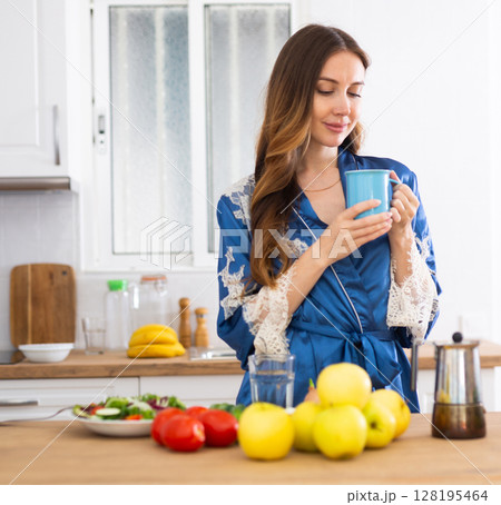 Young woman in blue dressing gown enjoying morning coffee in kitchen Young woman in blue dressing gown enjoying morning coffee in kitchen 128195464