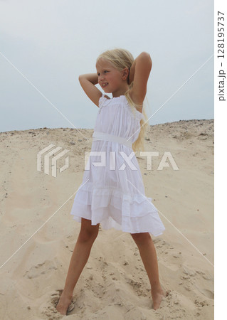 Cheerful young blond child girl in white dress standing on sand outdoors Cheerful young blond child girl in white dress standing on sand outdoors 128195737