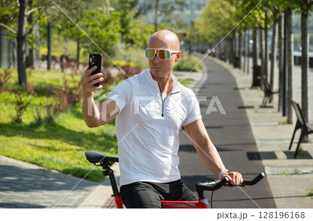 Handsome caucasian adult cyclist taking selfie or talk and video by smart phone in park. Outdoor activity. Handsome caucasian adult cyclist taking selfie or talk and video by smart phone in park. Outdoor activity. 128196168