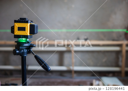 A laser level mounted on a tripod projects a green line onto a construction site. A laser level is essential for precise alignment and measurements in construction projects. 128196441