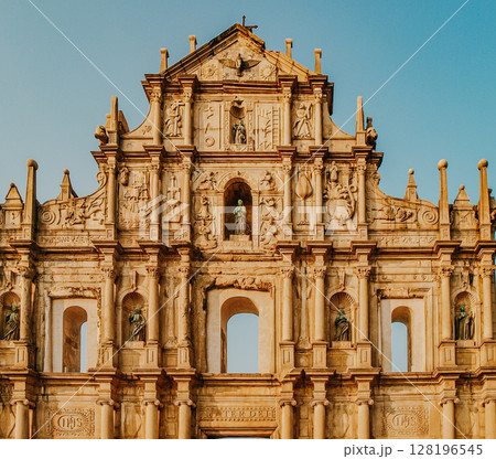 Historic Facade Of The Ruins Of St. Pauls In Macau: Ornate Stone Architecture With Religious Sculptures And Carvings Glowing In Warm Evening Light Against A Clear Sky 128196545