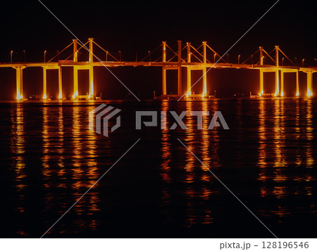 Illuminated Bridge In Hong Kong At Night: Glowing Golden Lights Reflecting On Calm Water Surface Beneath A Modern Suspension Bridge In A Coastal Urban Landscape 128196546