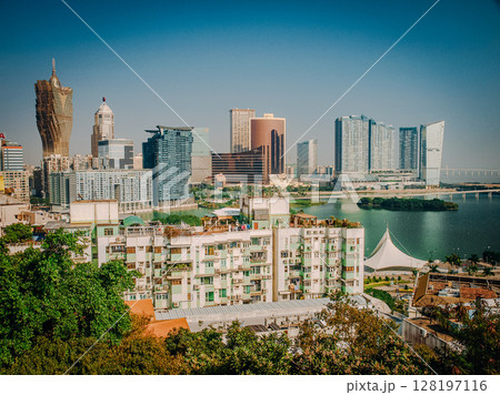 Urban Cityscape Of Macau From Above: Colorful Rooftops Of Historic District Blending Into High-Rise Buildings And Harbor Activity Across The Water On A Hazy Day 128197116