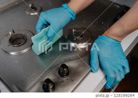 Cropped shot of male hand wearing blue gloves meticulously cleaning gas stove with sponge and detergent, ensuring hygiene and sanitation in kitchen environment. Concept of cleaning and household. Cropped shot of male hand wearing blue gloves meticulously cleaning gas stove with sponge and detergent, ensuring hygiene and sanitation in kitchen environment. Concept of cleaning and household. 128199264