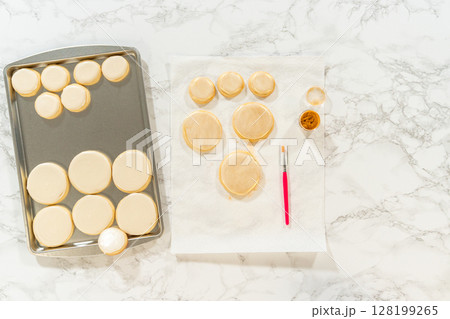 A baking sheet filled with round sugar cookies covered in smooth royal icing, ready for decorating. A brush, edible gold dust, and paper towels sit nearby for the final touches. 128199265