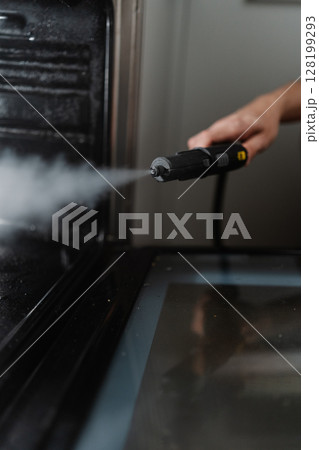 Vertical cropped shot of cleaner person sanitizing black gas oven interior with high-temperature steam, demonstrating kitchen cleaning hygiene process, close-up. Concept of cleanliness, housekeeping. 128199293