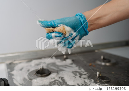 Cleaning service employee wearing protective gloves squeezing soapy water from sponge during cleaning dirty gas stove, to removing stubborn grime and grease. Concept of domestic work and household. 128199308