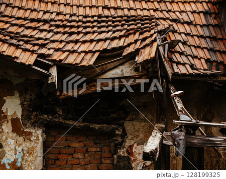 Collapsing Wooden House With A Broken Roof And Crumbling Walls: An Abandoned Rural Structure Overgrown With Greenery And Wildflowers In A Natural Landscape 128199325