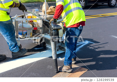 Workers are painting white road lines on an asphalt surface while traffic passes by under clear skies. Workers are painting white road lines on an asphalt surface while traffic passes by under clear skies. 128199539
