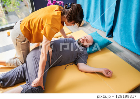Masseuse making thai yoga massage. Woman therapist doing traditional massage treatment, stretching male patient's body. Bald client lying on mat, enjoying hand massage in calm atmosphere. 128199817