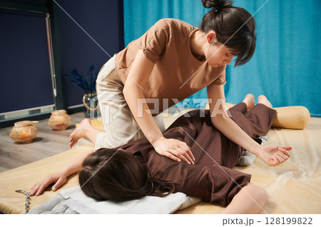 Masseuse making thai yoga massage. Woman therapist doing traditional massage treatment for female patient using elbow. Client lying on mat, meditating, enjoying hand massage in calm atmosphere. Masseuse making thai yoga massage. Woman therapist doing traditional massage treatment for female patient using elbow. Client lying on mat, meditating, enjoying hand massage in calm atmosphere. 128199822