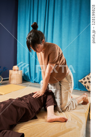 Masseuse making thai yoga massage. Woman therapist doing traditional massage treatment, stretching female patient's body. Client lying on mat, enjoying hand massage in calm atmosphere. Masseuse making thai yoga massage. Woman therapist doing traditional massage treatment, stretching female patient's body. Client lying on mat, enjoying hand massage in calm atmosphere. 128199828