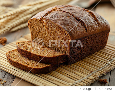 A freshly baked loaf of brown bread, rests on a bamboo mat alongside wheat stalks, highlighting its rustic appeal. 128202568