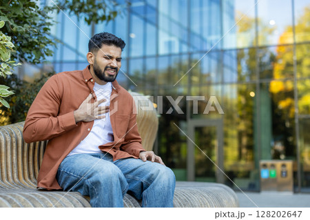 A man experiencing chest pain, sitting outdoors, likely having a health issue or heart attack, in a city setting. A man experiencing chest pain, sitting outdoors, likely having a health issue or heart attack, in a city setting. 128202647