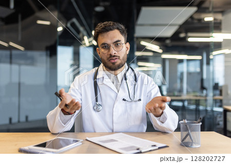 Latin American physician discussing medical topics while seated at an office desk with documents and tools. Depicts professional consultation, healthcare expertise, and effective communication Latin American physician discussing medical topics while seated at an office desk with documents and tools. Depicts professional consultation, healthcare expertise, and effective communication 128202727