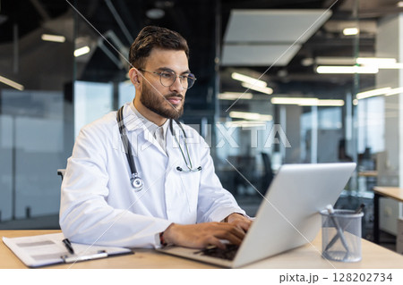 Young doctor works on his laptop in a well lit modern office. He wears a white coat and stethoscope while working at his desk Young doctor works on his laptop in a well lit modern office. He wears a white coat and stethoscope while working at his desk 128202734