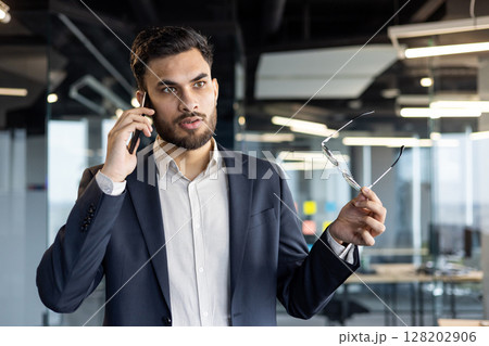 Businessman taking important phone call in office. Holds glasses while conversing, conveys confidence, communication, professionalism. Modern business environment. Tags: businessman, phone, office. 128202906