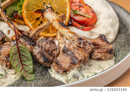 Grilled lamb chops served on a creamy herb sauce with roasted vegetables, flatbread, and a slice of dried orange for garnish 128203198