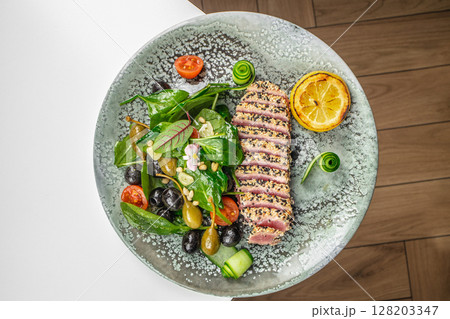 A beautifully plated dish featuring sesame-crusted seared tuna slices served with a fresh spinach and olive salad, cherry tomatoes, pine nuts, cucumber spirals, and grilled lemon A beautifully plated dish featuring sesame-crusted seared tuna slices served with a fresh spinach and olive salad, cherry tomatoes, pine nuts, cucumber spirals, and grilled lemon 128203347