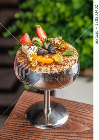 Luxury tiramisu in a silver coupe topped with berries, pistachios, mint, and edible gold 128203402