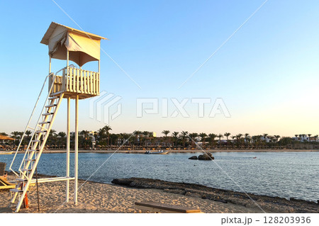 A lifeguard tower stands on a sandy beach by the Red Sea in Egypt. The scene captures a clear summer sky and calm waters, with palm trees in the background. A lifeguard tower stands on a sandy beach by the Red Sea in Egypt. The scene captures a clear summer sky and calm waters, with palm trees in the background. 128203936