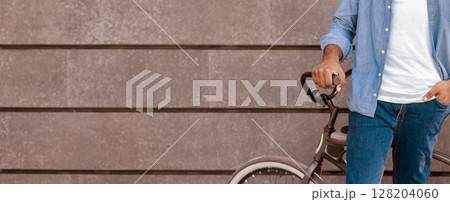 Closeup of unrecognizable african man standing near his bike, holding handlebar, posing over grey urban wall Closeup of unrecognizable african man standing near his bike, holding handlebar, posing over grey urban wall 128204060