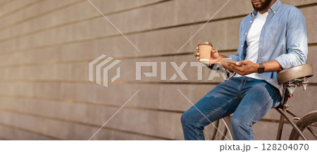 Black millennial guy with smarthone and takeaway coffee sitting on bicycle, relaxing outdoors next to gray urban wall. copy space 128204070