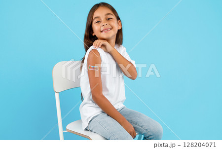Vaccination Of Children. Happy Vaccinated Kid Girl Showing Arm With Adhesive Bandage After Vaccine Injection Sitting On Blue Studio Background. Kids And Covid-19 Prevention, Antiviral Immunization Vaccination Of Children. Happy Vaccinated Kid Girl Showing Arm With Adhesive Bandage After Vaccine Injection Sitting On Blue Studio Background. Kids And Covid-19 Prevention, Antiviral Immunization 128204071