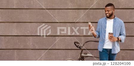 Black guy standing next to bicycle, texting on smartphone and drinking takeaway coffee outdoors, copy space Black guy standing next to bicycle, texting on smartphone and drinking takeaway coffee outdoors, copy space 128204100